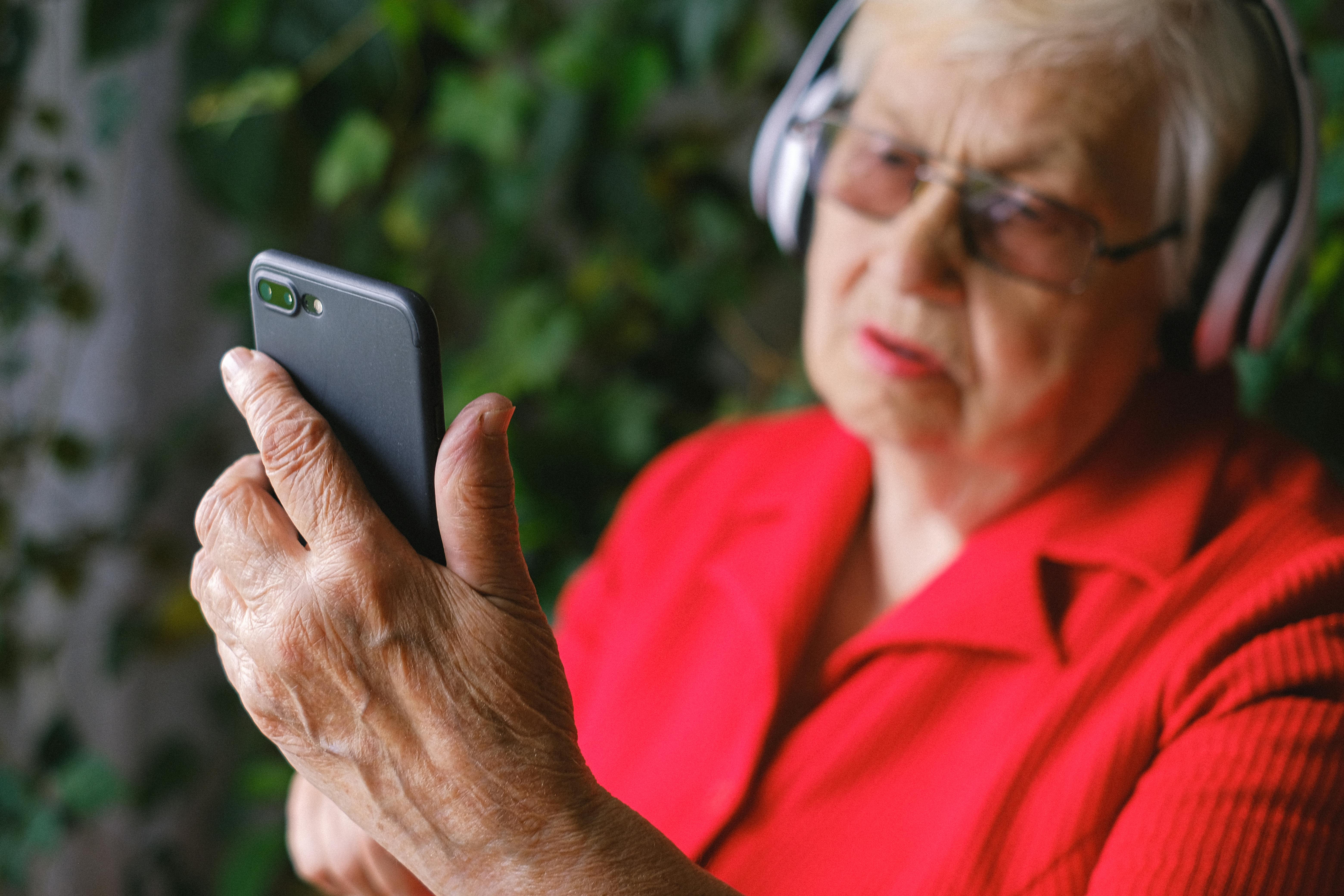 A person listening to music on a smartphone wearing headphones. The person has a red blouse on, glasses and grey hair. 