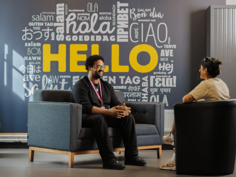 Two people sat in conversation at a One Manchester meeting. The wall behind features the words Hello in multiple languages.