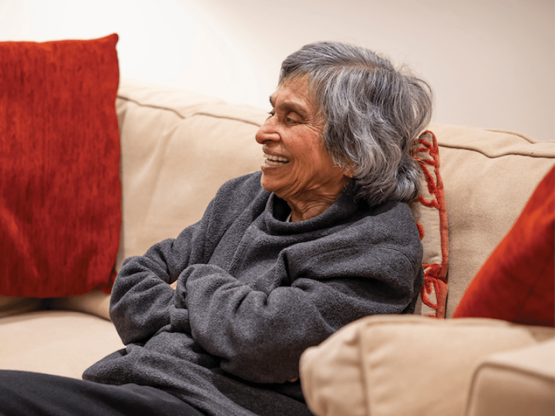 A woman sat on a sofa. She's wearing a jumper and has grey hair.