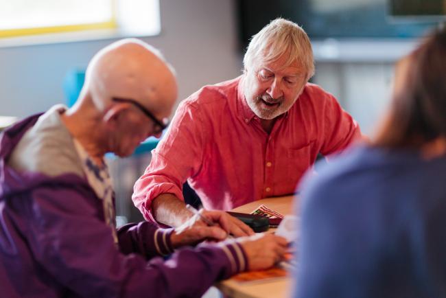 Two men sat at a table, one is writing and the other is talking. 